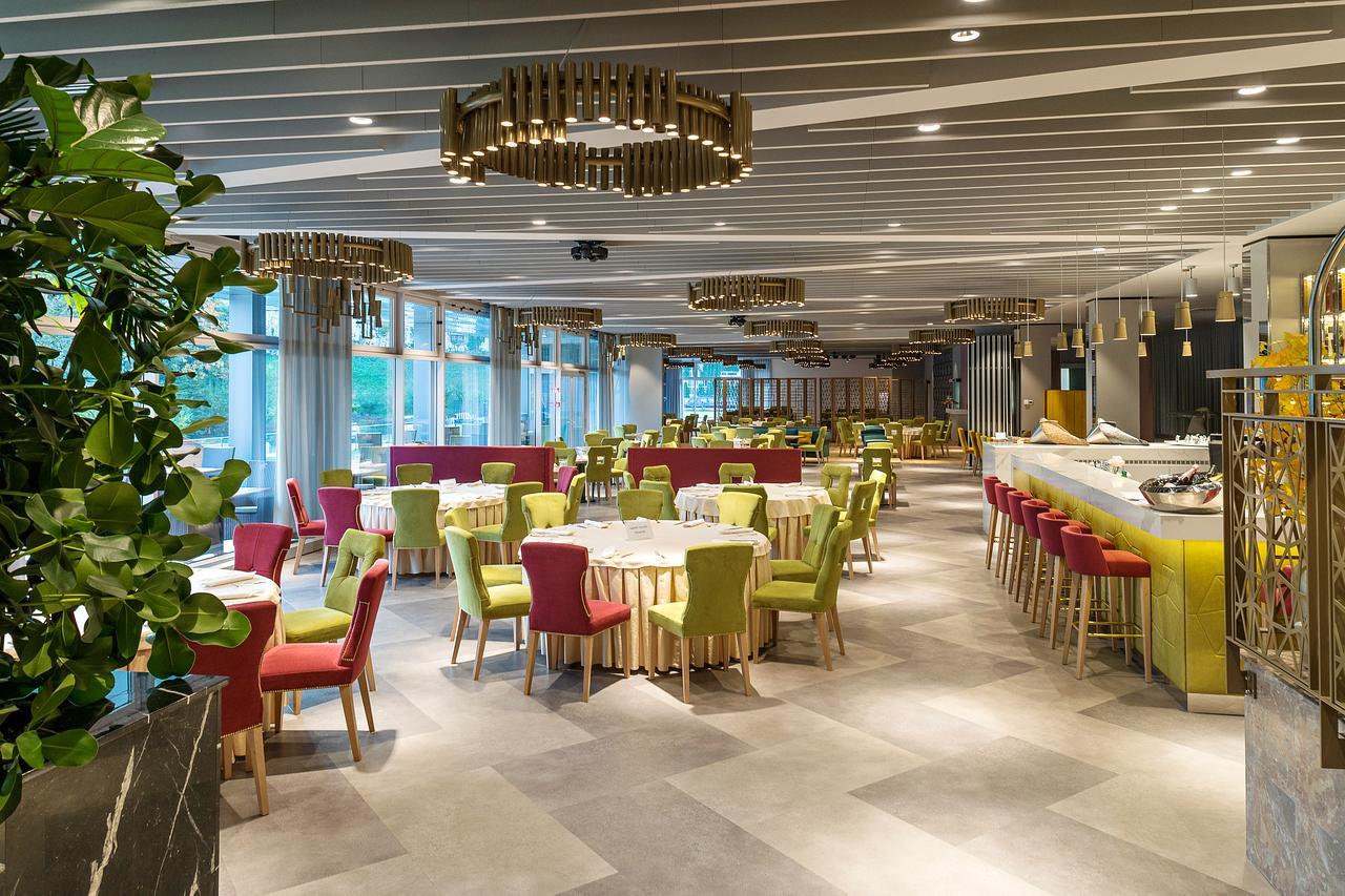Bright, modern restaurant dining area with colorful upholstered chairs around round tables and a bar with yellow panels on the right.