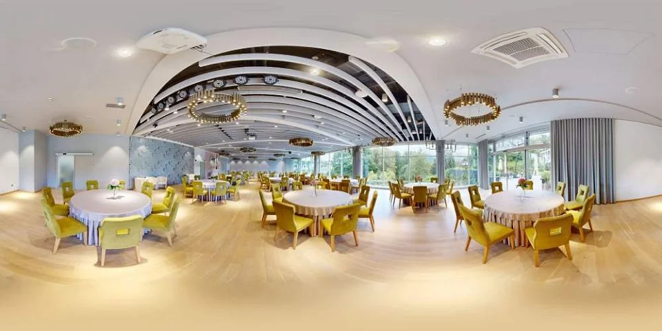 Spacious banquet hall with round tables, white tablecloths, and yellow chairs, bright with large windows and gold chandeliers.