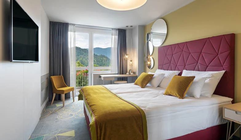 Hotel room with a large bed, mustard‑yellow runner and pillows, purple upholstered headboard, and circular mirrors on the wall.