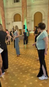 Group of people in a ballroom standing apart, likely warming up or practicing a dance routine in a grand room with ornate columns and arches.
