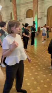 People dancing in a grand hall with marble columns and gold curtains; participants stand in a line, practicing a routine on a checkered wooden floor.