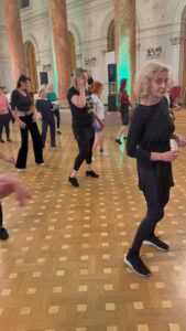 Group of people dancing in a grand ballroom with marble columns and green uplighting, practicing steps on a polished parquet floor.