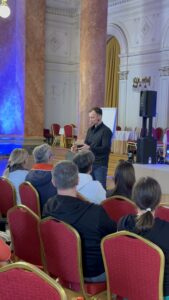 Male speaker in a black shirt giving a talk to an audience in a grand ballroom with marble columns and ornate white walls.