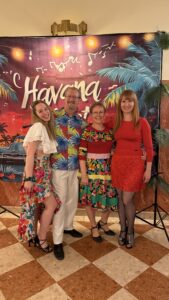 Group of four adults posing in tropical outfits in front of a Havana-themed backdrop with palm leaves and string lights.