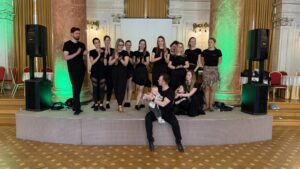 Group of performers in black on a stage posing for a photo, with green uplighting and two large marble columns; a man sits front center holding a baby while others applaud nearby.