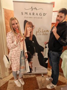 Two people stand in front of a Smarag d plesni studio banner, each licking an orange popsicle while holding bags.