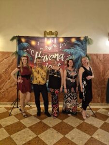 Five adults pose for a group photo in front of a tropical Havana Nights backdrop at an indoor event.