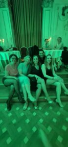Group of five women seated on steps, smiling at a social event with candlelit tables in the background