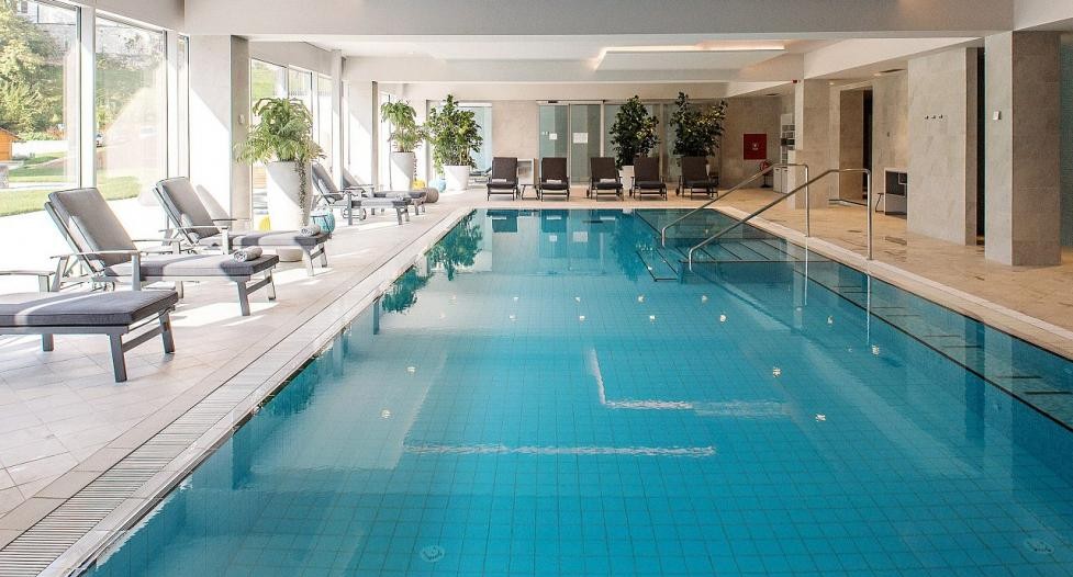 Indoor swimming pool area with blue water, lounge chairs along the left, large windows, and potted plants in the background.
