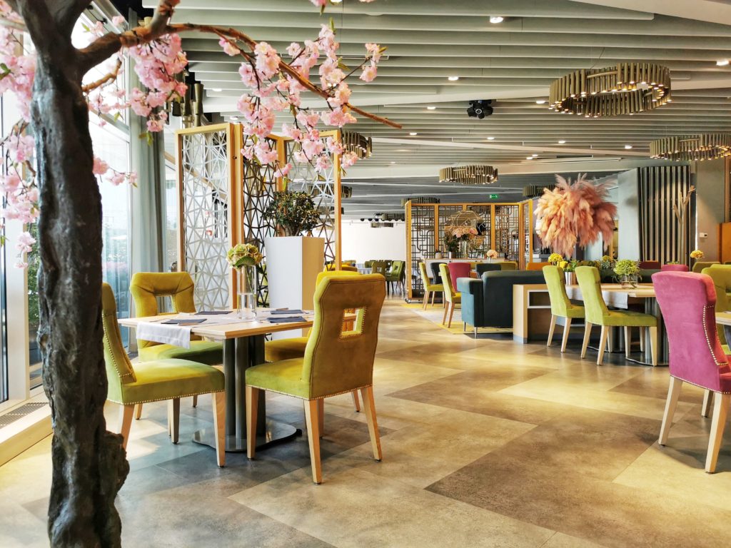 Bright restaurant dining area with colorful upholstered chairs (yellow, green, pink) around light wooden tables; a flowering tree branches across the foreground; decorative partitions and warm pendant lighting create a cozy, stylish atmosphere.