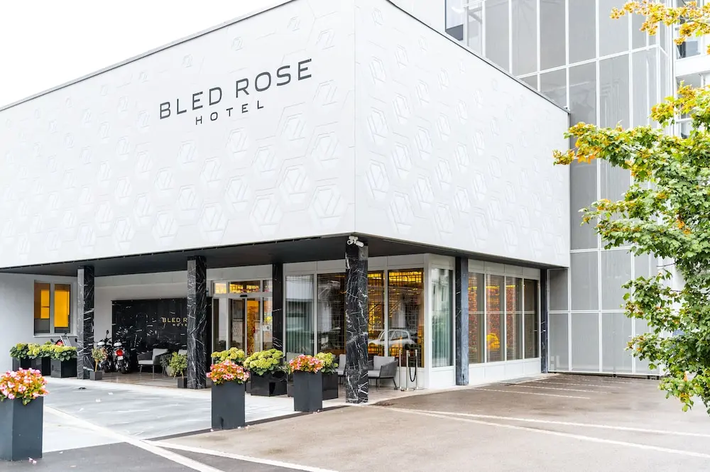 Exterior view of the Bled Rose Hotel with a white patterned facade, glass entry, and potted flowers along the sidewalk