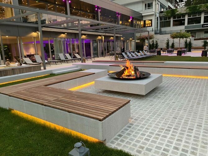 Modern outdoor courtyard with a central square fire pit on a raised concrete pedestal, surrounded by wooden benches, gray tiled paving, and lounge chairs along a glass-walled building with warm ambient lighting.